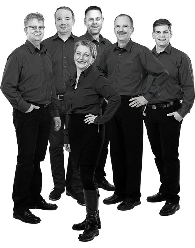 Sechs Personen in schwarzer Arbeitskleidung stehen zusammen, eine Frau vorne mit Hand in der Hüfte, fünf Männer im Hintergrund, alle in Hemden und Hosen.