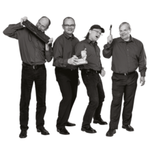 Vier Männer in dunklen Hemden und Hosen stehen nebeneinander, einer hält eine Tastatur, ein anderer einen Schraubendreher, zwei zeigen Daumen hoch.